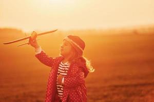 Illuminated by orange colored sunlight. Cute little girl have fun with toy plane on the beautiful field at daytime photo