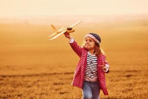 Illuminated by orange colored sunlight. Cute little girl have fun with toy plane on the beautiful field at daytime photo