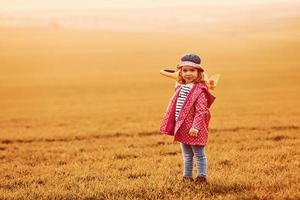 Illuminated by orange colored sunlight. Cute little girl have fun with toy plane on the beautiful field at daytime photo