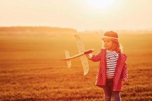 Illuminated by orange colored sunlight. Cute little girl have fun with toy plane on the beautiful field at daytime photo