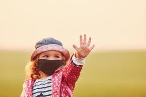 Showing hand. Portrait of cute little girl in black protective mask that standing outdoors photo