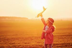 Illuminated by orange colored sunlight. Cute little girl have fun with toy plane on the beautiful field at daytime photo