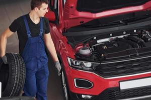 Worker in black and blue uniform holds car wheel and walks with in near red automobile photo