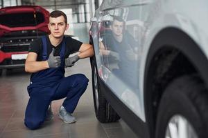 Guy in blue uniform sitting near fixed white car and shows thumb up photo