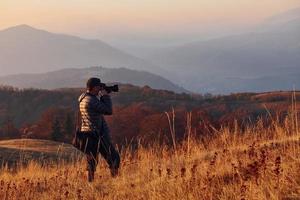 fotógrafo masculino de pie y trabajando en el majestuoso paisaje de árboles y montañas de otoño en el horizonte foto