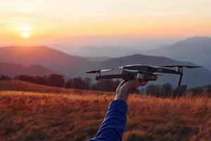 Man standing in the autumn field and holds drone in hand. Beautiful sunrise. Majestic landscape of mountains far away photo