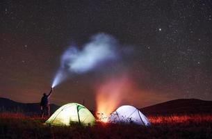 el hombre sostiene un dispositivo con haz de luz. dos carpas iluminadas con fogata bajo las estrellas en las montañas por la noche foto