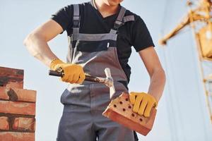 Holding brick and using hammer. Construction worker in uniform and safety equipment have job on building photo