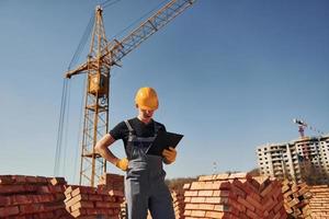 Holds notepad with documents. Construction worker in uniform and safety equipment have job on building photo