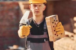 Portrait of construction worker in uniform and safety equipment that standing on building and holding brick and tool photo