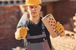 Portrait of construction worker in uniform and safety equipment that standing on building and holding brick and tool photo