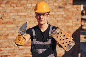 Portrait of construction worker in uniform and safety equipment that standing on building and holding brick and tool photo