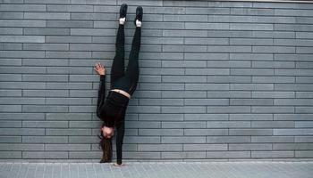 joven deportista con ropa deportiva negra haciendo ejercicios de parada de manos duros al aire libre cerca de la pared gris foto