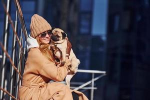 Woman in warm clothes sitting and have fun with her little pug dog near business building that on background photo