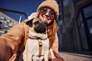 Woman in warm clothes make selfie with her little pug dog near business building that on background photo