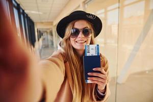 joven pasajera con ropa abrigada que muestra boletos en la sala del aeropuerto foto