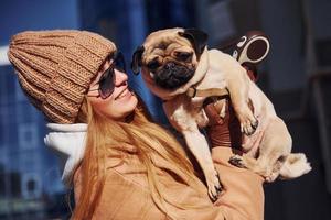 Woman in warm clothes have holding her little pug dog on hands near business building that on background photo