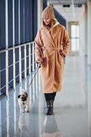 Woman in warm clothes walking with her little pug dog indoors in the hall photo