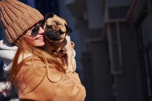 Woman in warm clothes have holding her little pug dog on hands near business building that on background photo