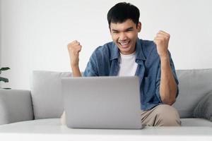 Happy excited Asian man looking at laptop, man receiving good news using computer at home. photo