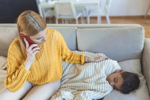 joven madre, sosteniendo a su pequeño niño enfermo, acostados juntos en el sofá. mamá llamando al médico por su hijo enfermo foto