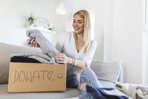 Happy blond young woman sit on couch stuck clothes in donation box at home, caring biracial female volunteer put apparel in carton package, donate to needy people, reuse, recycle concept photo