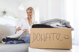 Woman packing clothes into donation box in living room. Girl puts in a box with donations items. Volunteering. Woman participating at charity and holding donation box photo