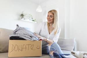 Young woman sitting on couch preparing parcel for sending to needy human. Girl with big kind heart puts used clothes new wear in donation box, concept of caring about homeless people photo