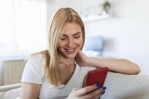 Young woman sit relax on sofa in living room browsing surfing wireless internet on smartphone, millennial girl rest on couch at home message text on modern cellphone, shopping online via website photo
