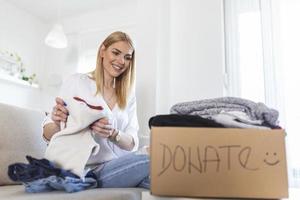 Young woman with donation box at home. Donation box for poor with clothing in female hands. Woman donates clothing to shelter. Cheerful woman holds box of donated clothing photo