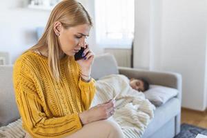 mujer joven que controla la temperatura con la mano del pequeño hijo enfermo. madre llamando a un médico para su niño enfermo niño enfermo acostado en la cama bajo una manta con una mujer controlando la fiebre en la frente con la mano. foto