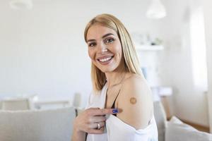 Beautiful woman looking happy after getting vaccine. Female with band-aid on her arm after receiving covid-19 vaccination sitting at home photo