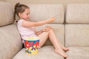 cute joyful girl sits at home on the sofa with a huge glass of popcorn and switches TV channels with a remote control photo