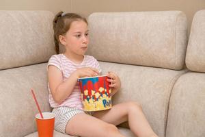 cute girl with a pigtail sits at home on the couch watching a movie with enthusiasm, eating popcorn from a paper cup and drinking cola photo