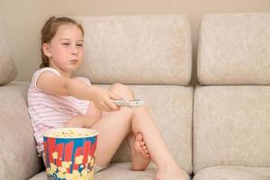 concentrated, squinted look of a little girl sitting at home on the couch with a huge glass of popcorn and switching channels on the TV with a remote control photo