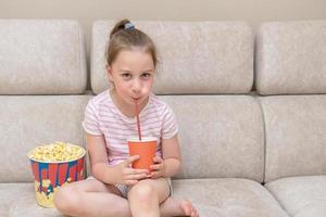 cute little girl sits on the couch at home and drinks a cocktail from a straw, next to it is a huge glass of popcorn photo