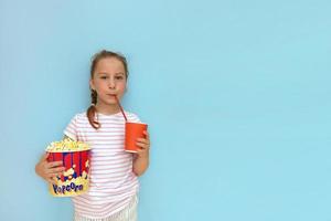 girl in a striped T-shirt holds a large glass of popcorn and drinks from a glass with a straw on a blue background with copy space photo