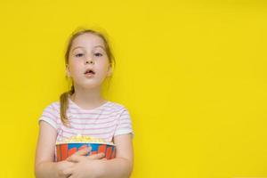 cute little girl with a lost front milk tooth wants to say something, holds a glass of popcorn with both hands, on a yellow background with copy space photo