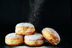 el azúcar en polvo se vierte sobre las donas. postre judío tradicional sufganiyot sobre fondo negro. cocinar berlineses fritos. foto