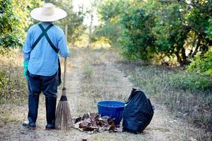 la vista posterior del jardinero usa sombrero, sostiene un palo de escoba, se encuentra al lado de una bolsa de basura negra y una canasta en el jardín. concepto, barrer las hojas secas para hacer compost. proteccion Ambiental foto