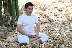 Portrait of Buddhist Asian man in white custom  doing meditation in peaceful nature in forest. Close eyes, make concentration , feel free and calm. Concept for make merit, pure mind, intelligence. photo