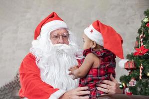 Santa Claus is lifting happy little toddler baby girl up and sitting on his lap with fully decorated christmas tree on the back for season celebration concept photo