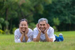 Portrait of senior Asian couple lying down on the grass in the public park looking at the camera with good mental health for longevity and happy marriage photo