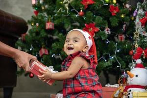 African American baby is happily smiling while receiving little gift box from parents while dressing in christmas dress and santa hat with christmas tree on the back for season celebration concept photo