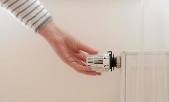 mujer regula la temperatura en casa con el termostato de calefacción para ahorrar energía, primer plano con la mano. válvula de control de temperatura en el radiador. calentador blanco en la pared con cortina en apartamento moderno. foto