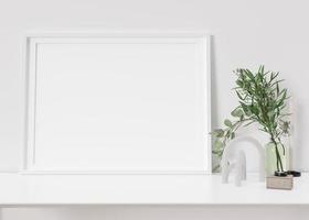 Empty horizontal picture frame standing on white shelf. Frame mock up. Copy space for picture, poster. Template for your artwork. Close up view. Plant in vase, home accessories, sculpture. 3D render. photo