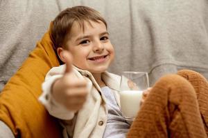 Little adorable boy sitting on the couch at home and drinking milk. Fresh milk in glass, dairy healthy drink. Healthcare, source of calcium, lactose. Preschool child with casual clothing. Thumbs up. photo