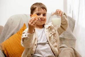 Little adorable boy sitting on the couch at home, drinking milk with cookie. Fresh milk in glass, dairy healthy drink. Healthcare, source of calcium, lactose. Preschool child with casual clothing. photo