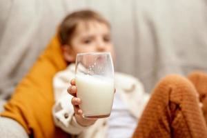 Little adorable boy sitting on the couch at home and drinking milk. Fresh milk in glass, dairy healthy drink. Healthcare, source of calcium, lactose. Preschool child with casual clothing. photo