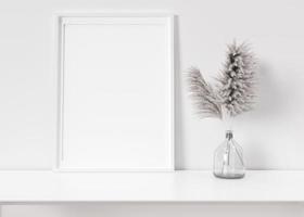 Empty vertical picture frame standing on white shelf. Frame mock up. Copy space for picture, poster. Template for your artwork. Close up view. Pampas grass in vase. 3D rendering. photo